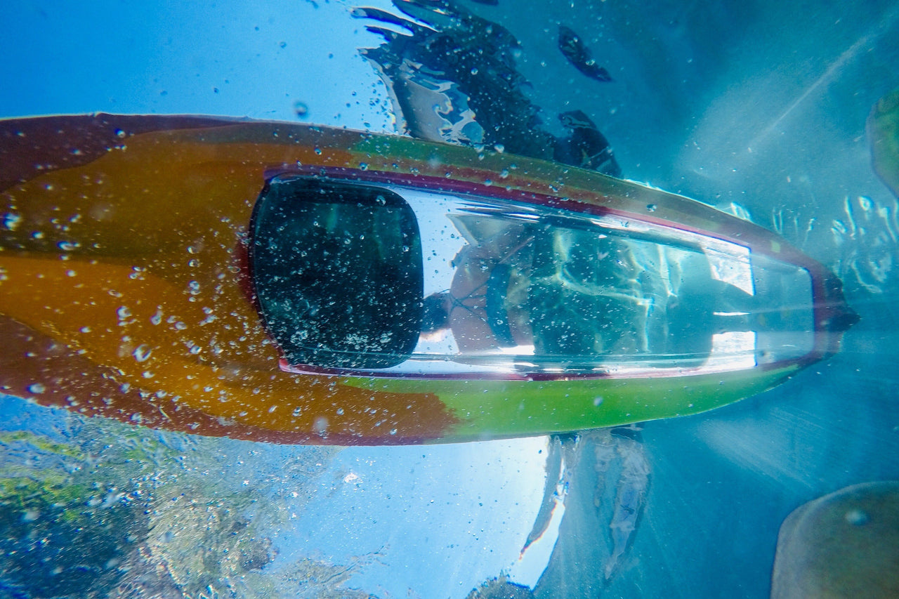 An underwater close-up of a transparent kayak with a person inside, showing blue water, bubbles, and reflections on the clear hull.